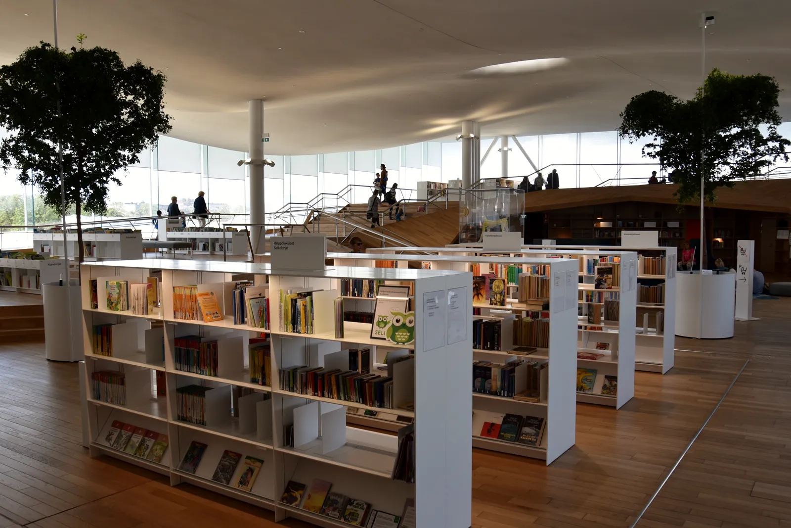 Lesesaal der Zentralbibliothek Oodi in Helsinki mit weißen Bücherregalen, Bäumen und geschwungener Decke